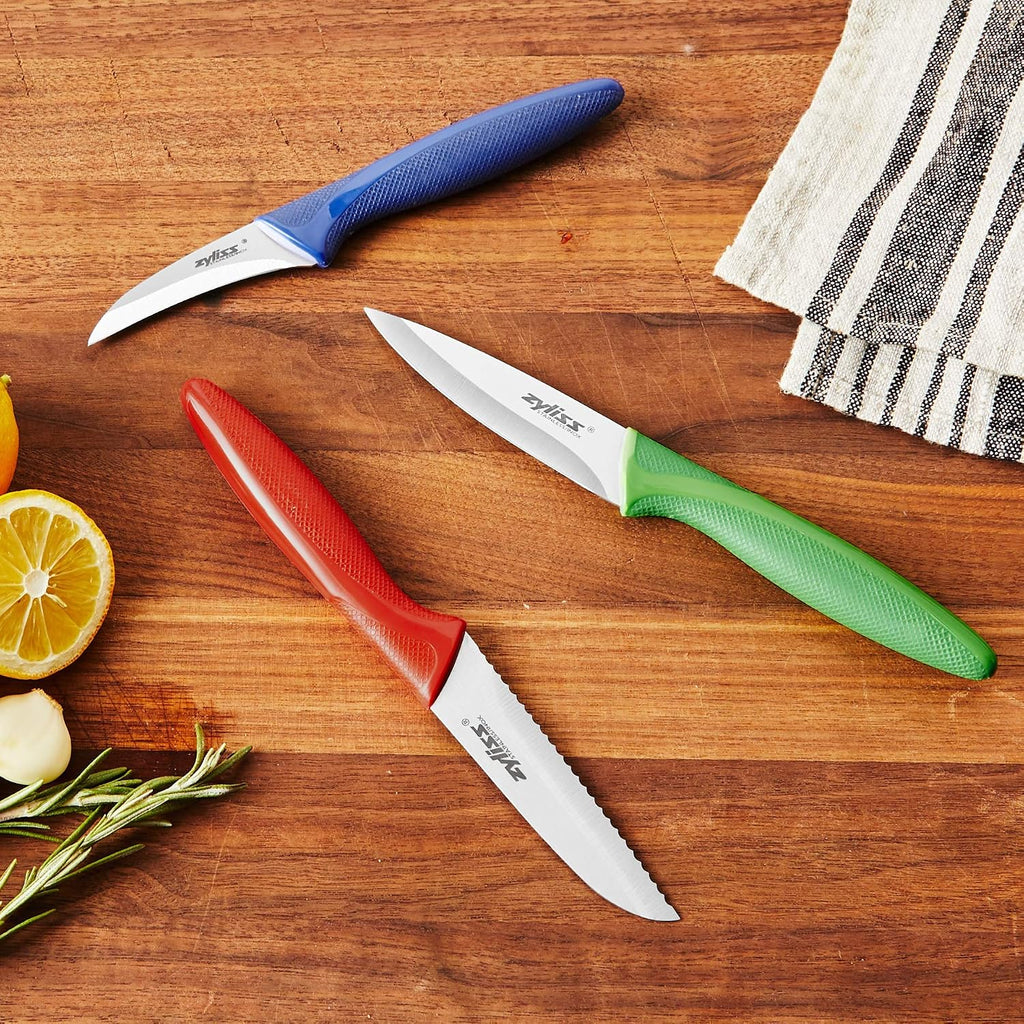 Three small kitchen knives with textured handles in blue, red, and green, arranged on a wooden cutting board with lemon, garlic, rosemary, and a striped cloth.